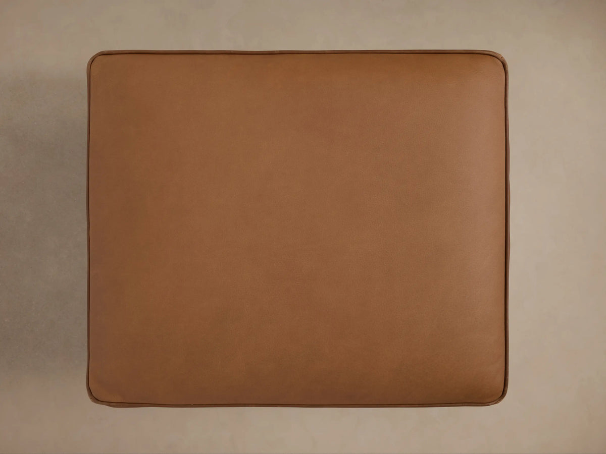 Angled view of a cognac leather Milly Leather Ottoman, featuring piped cushions, a natural wood frame, and turned legs.