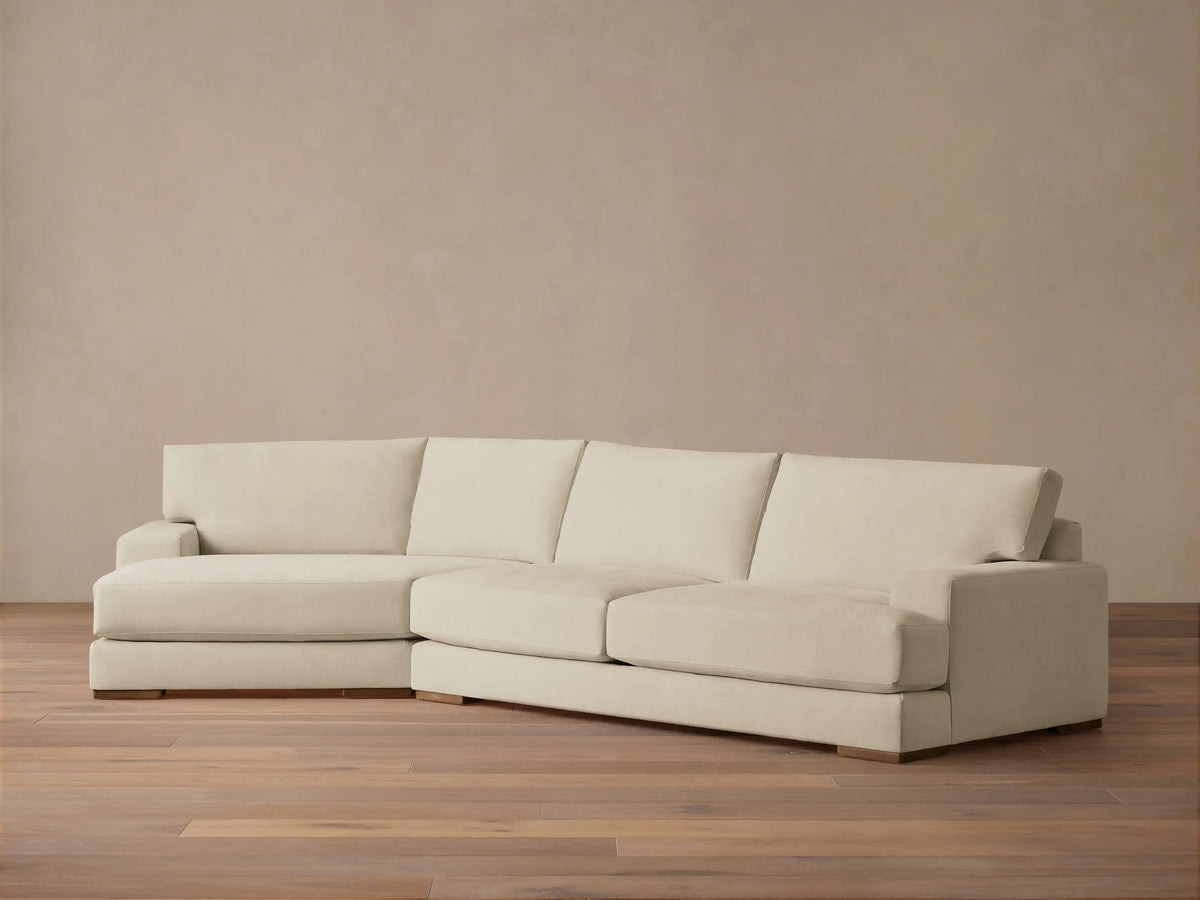 A beige Norwalk Two-Piece Cuddle Chaise Sectional on a wooden floor against a plain light-colored wall, featuring a chaise on the left.