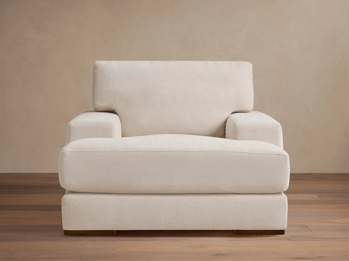 Norwalk Chair in off-white fabric with a plush cushion and backrest. It has wide arms and wooden legs, set on a wooden floor.