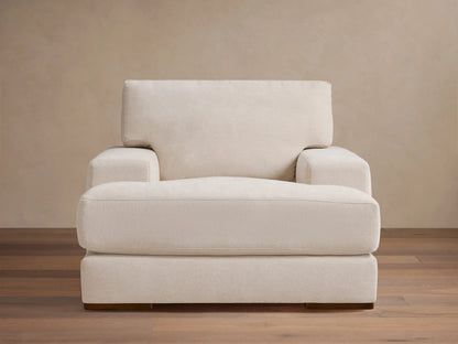 Norwalk Chair in off-white fabric with a plush cushion and backrest. It has wide arms and wooden legs, set on a wooden floor.