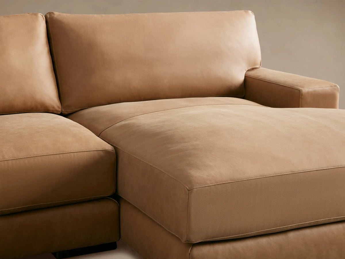 The Norwalk Leather Two-Piece Chaise Sectional in tan leather, with three seat cushions and an extended chaise on the right, sits on a hardwood floor.
