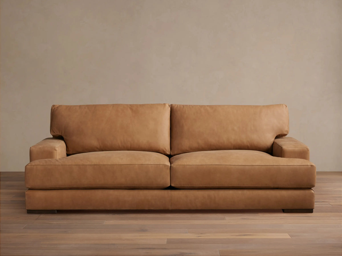 Tan leather Norwalk Sofa with two backrest cushions and sleek armrests placed on a wooden floor against a neutral wall.