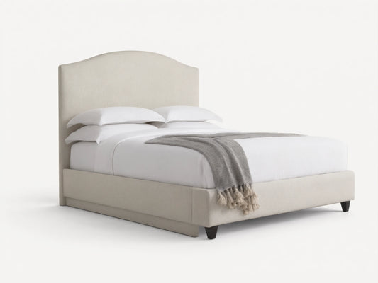 Omani Bed with cream upholstered arched headboard, white bedding, and a grey throw blanket with tassels, in a neutral room.