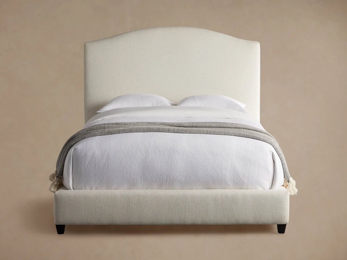 Omani Bed with cream upholstered arched headboard, white bedding, and a grey throw blanket with tassels, in a neutral room.
