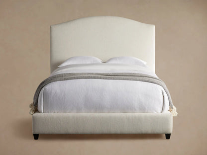 Omani Bed with cream upholstered arched headboard, white bedding, and a grey throw blanket with tassels, in a neutral room.