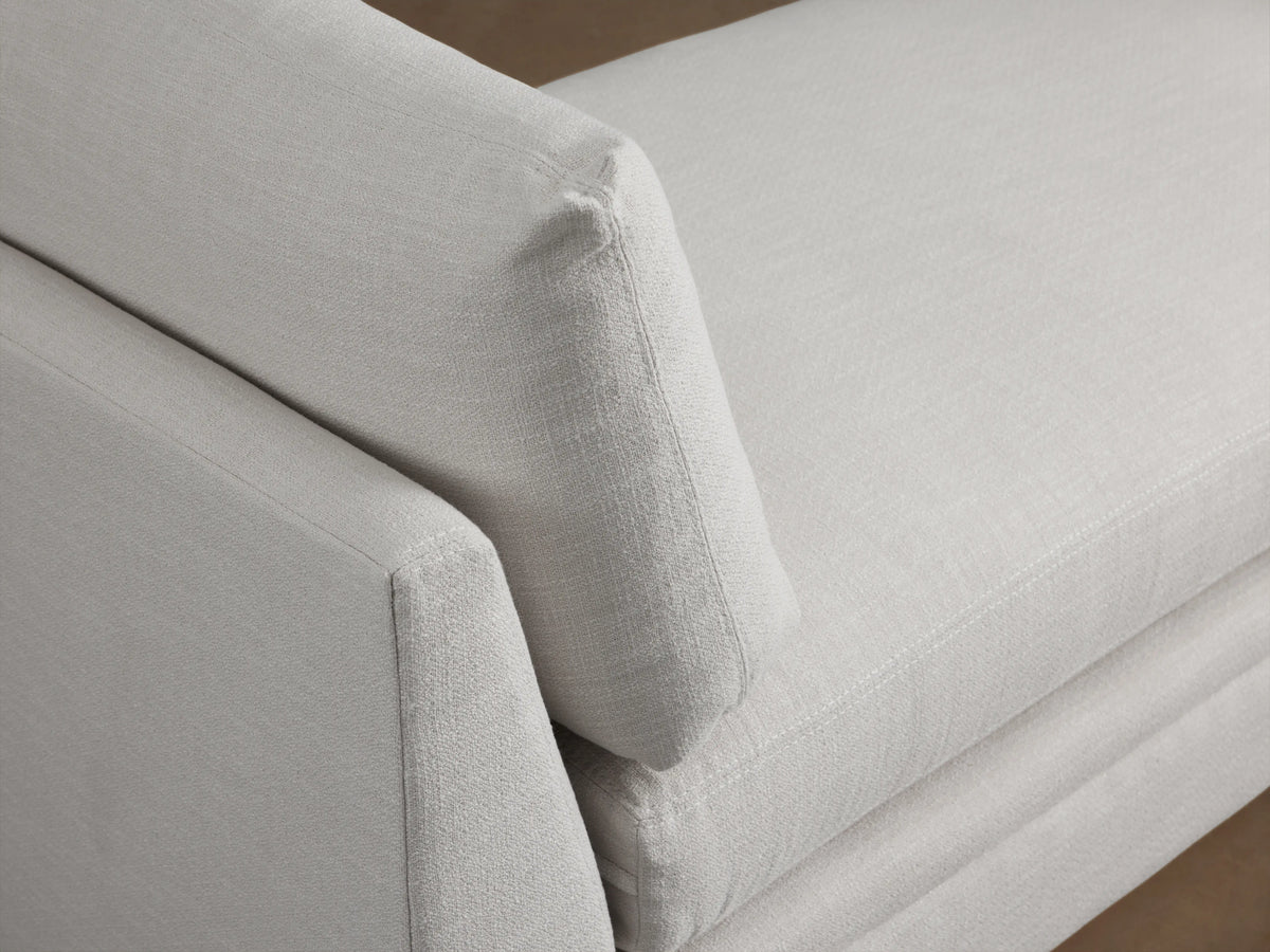 Pavo Daybed in cream fabric with rectangular shape. Features two large cushions at either end, supported by low-profile armrests, on wooden flooring against a neutral wall.