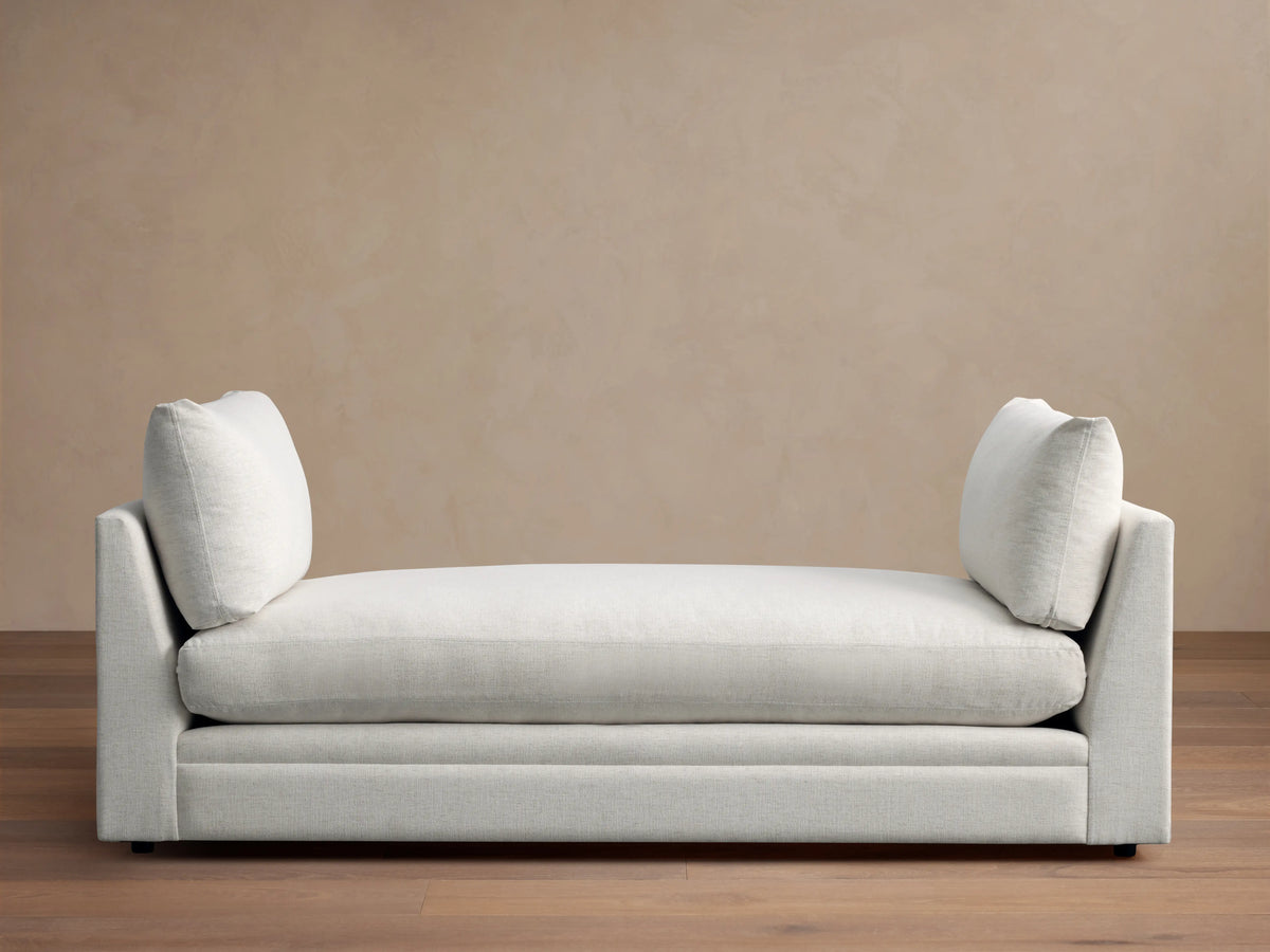Cream-colored Pavo Daybed with plush cushions on either end, set against a neutral wall on a wooden floor.