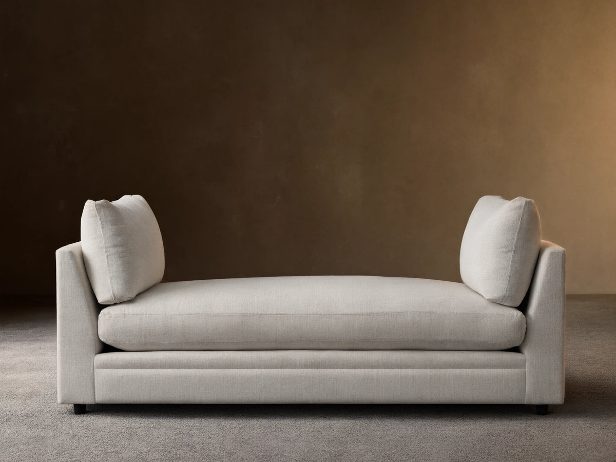 Cream-colored Pavo Daybed with plush cushions on either end, set against a neutral wall on a wooden floor.