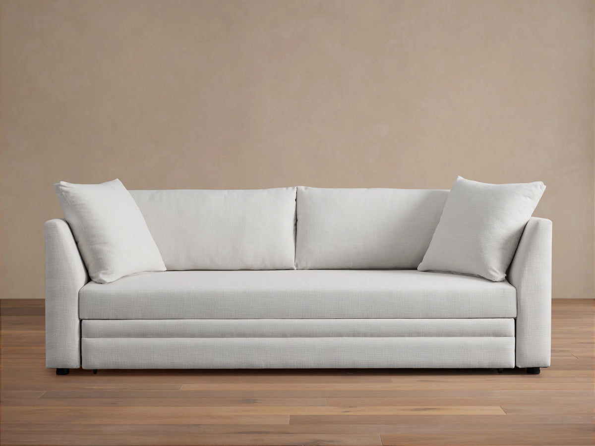 Pavo Trundle Sleeper Sofa in cream fabric, features a sleek design with straight arms and three large seat cushions. Set in a minimalist room.