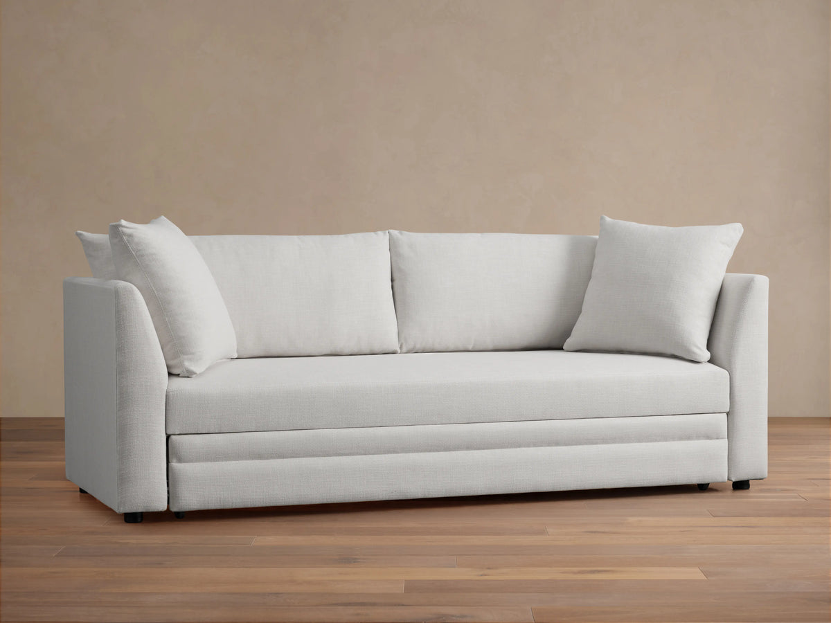 Pavo Trundle Sleeper Sofa in cream fabric, features a sleek design with straight arms and three large seat cushions. Set in a minimalist room.