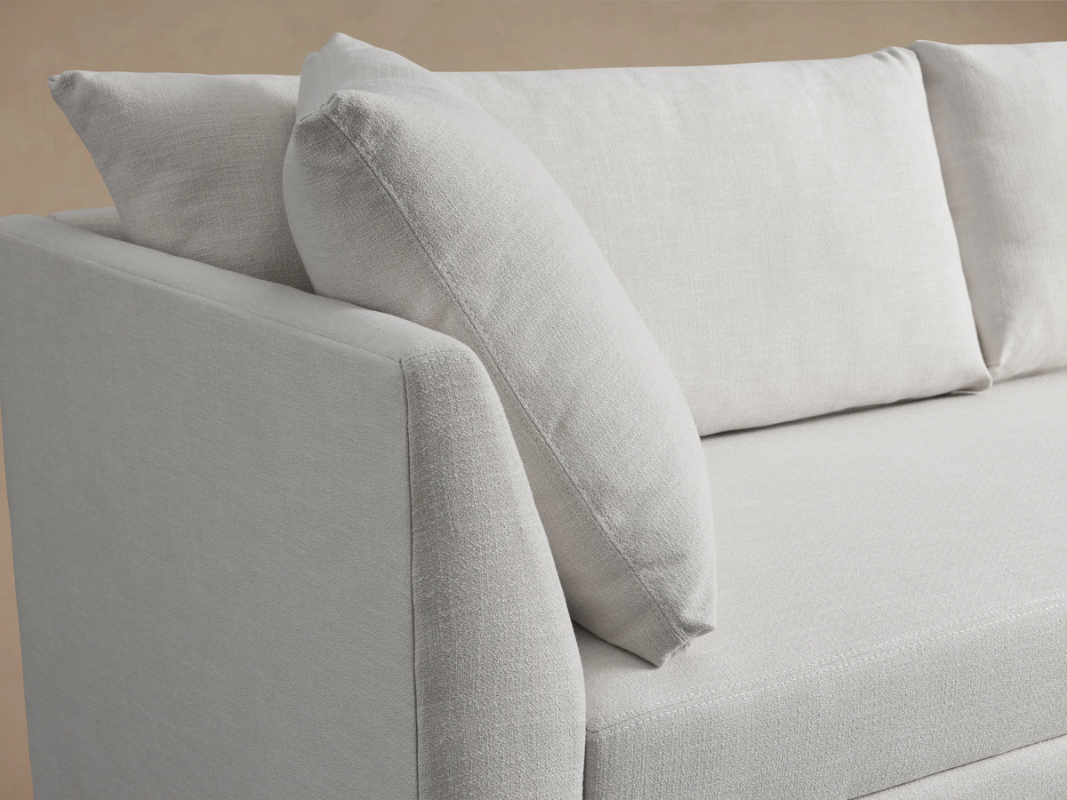 Pavo Trundle Sleeper Sofa in cream fabric with three back cushions, two side cushions, and a streamlined design on a wooden floor.