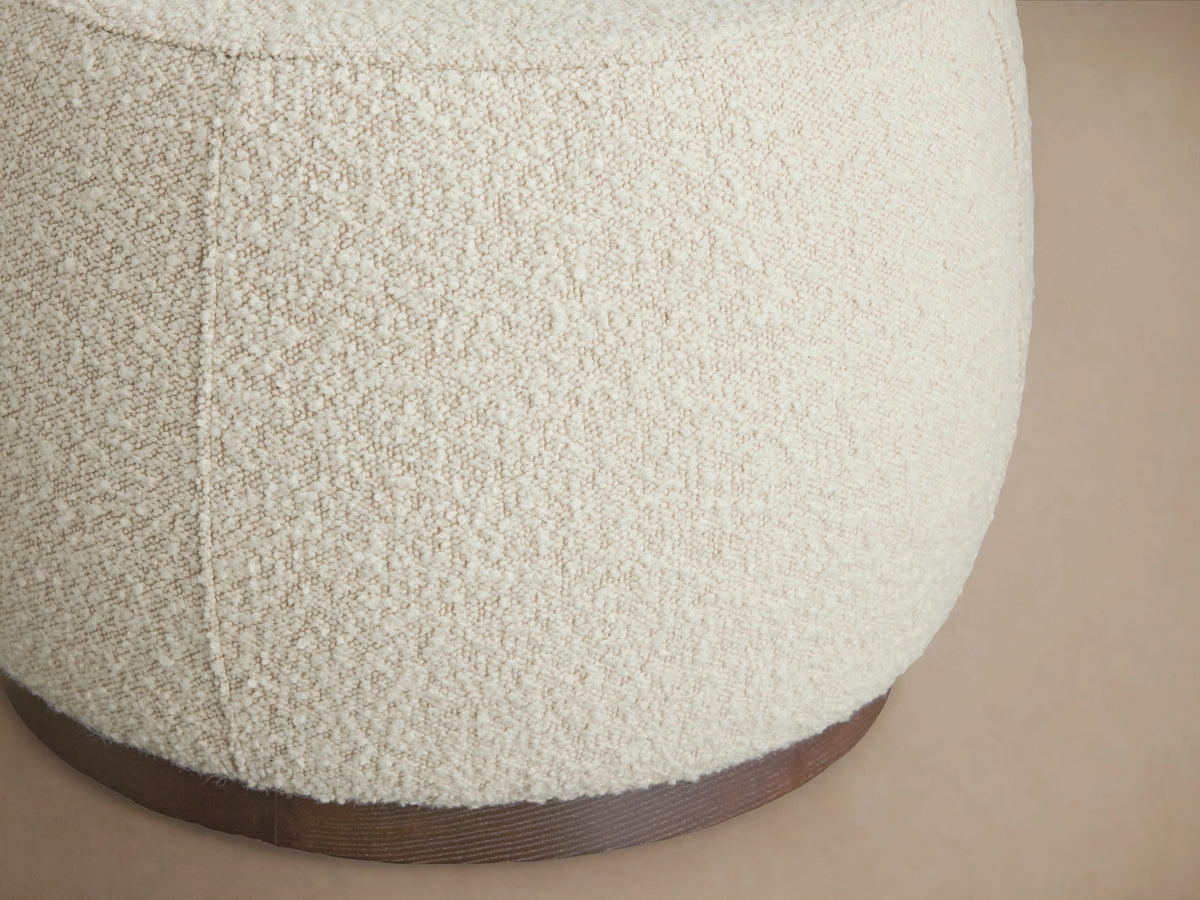 Round beige upholstered Ryles Plinth Ottoman with textured fabric, featuring a sturdy dark wood base. Close-up angle.