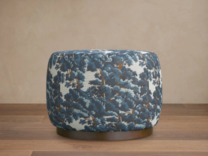 Ryles Plinth Ottoman in Oaks Pacific with a rounded shape, featuring a textured, blue and white tree pattern fabric, on a dark base, against a neutral wall.