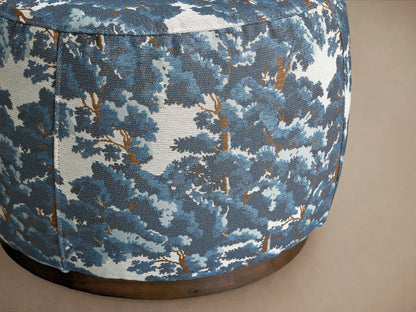 Ryles Plinth Ottoman in Oaks Pacific with a rounded shape, featuring a textured, blue and white tree pattern fabric, on a dark base, against a neutral wall.