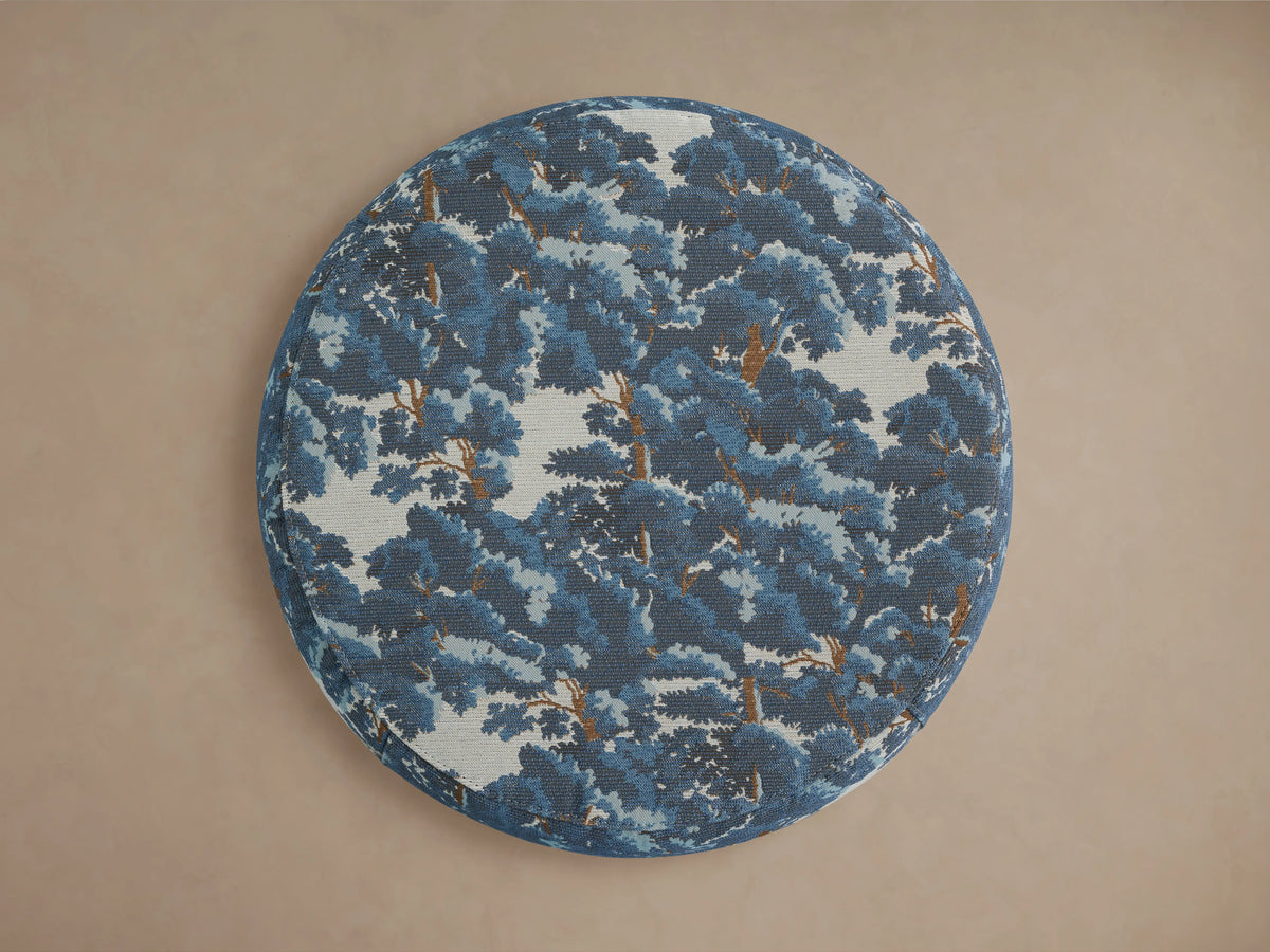 Ryles Plinth Ottoman in Oaks Pacific fabric with blue and white tree pattern, round shape, and wooden base on a beige background.