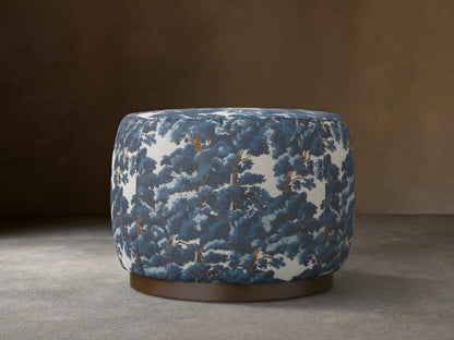 Ryles Plinth Ottoman in Oaks Pacific with a rounded shape, featuring a textured, blue and white tree pattern fabric, on a dark base, against a neutral wall.