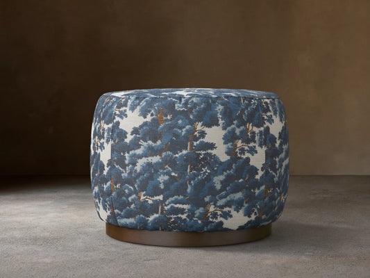 Ryles Plinth Ottoman in Oaks Pacific with a rounded shape, featuring a textured, blue and white tree pattern fabric, on a dark base, against a neutral wall.