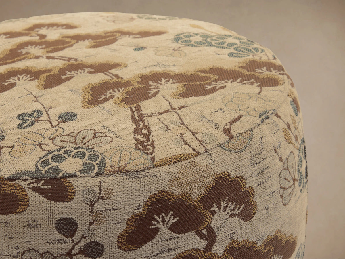 Ryles Ottoman upholstered in beige fabric with brown pine tree and blue floral pattern, sitting on a wooden floor.