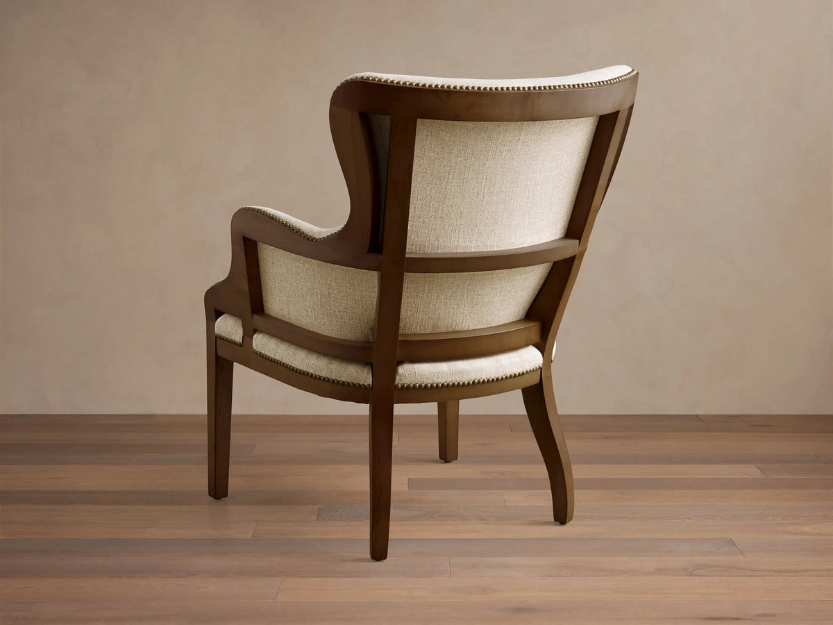 Rear view of the Tilly Chair, showing its dark wood frame, light beige woven upholstery, and brass nailhead detailing.