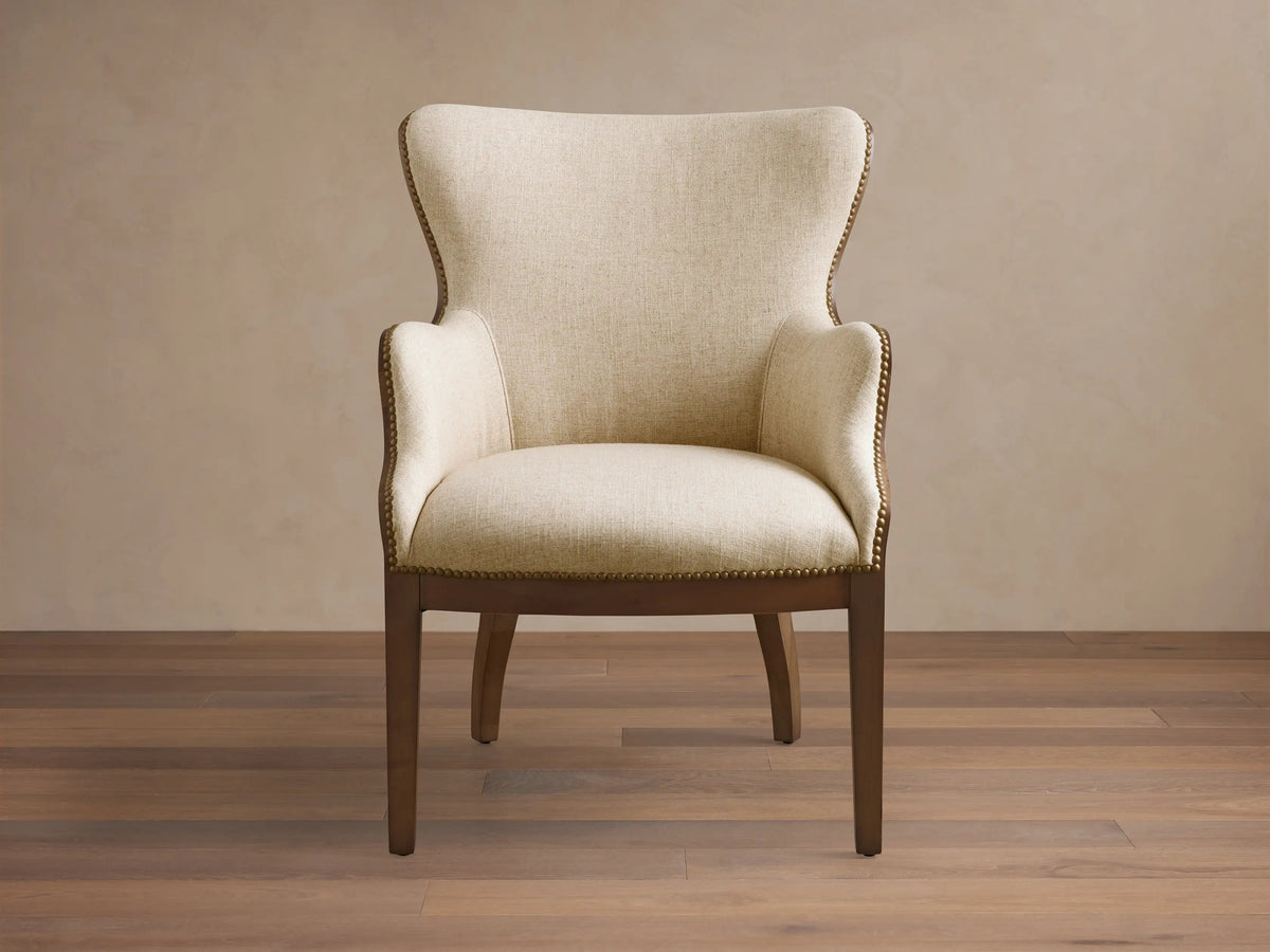 Front view of the light beige Tilly Chair, featuring a dark wood frame and delicate brass nailhead detailing.