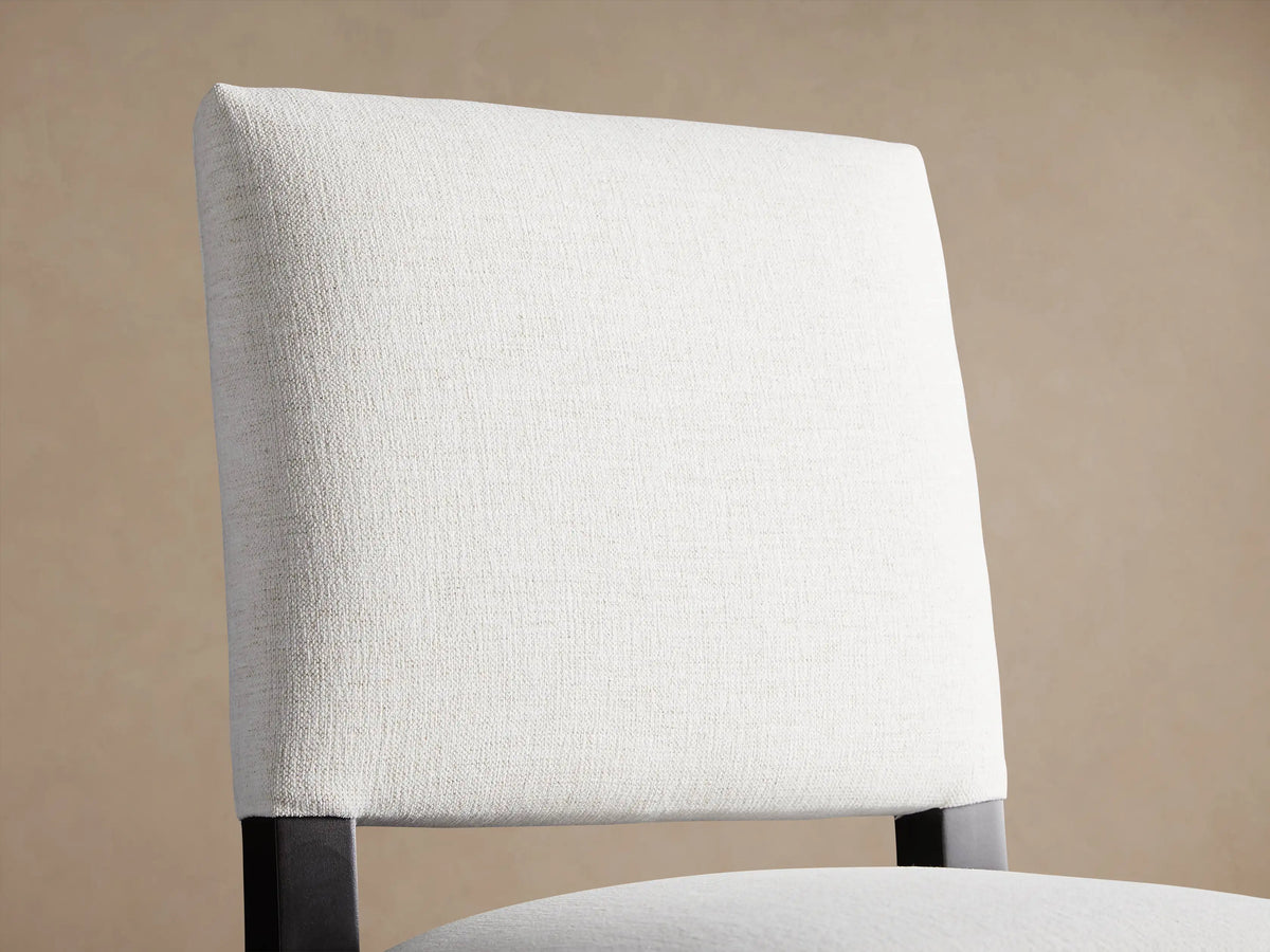 Light grey upholstered Torino Stool with dark wood frame and splayed legs, shown frontally against a beige wall on a wood floor.