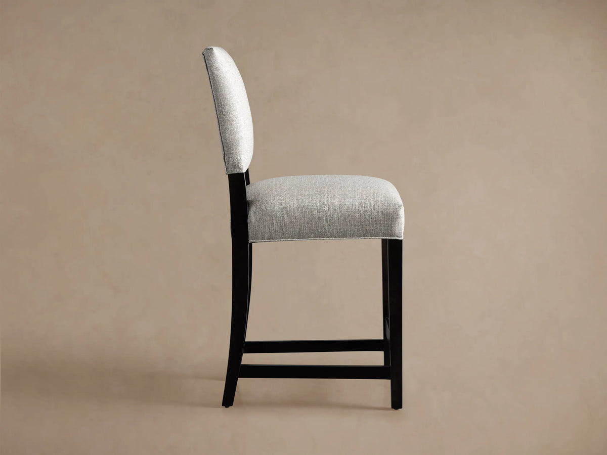 Torino Stool, back view. Light grey upholstered seat and backrest, dark wood tapered & splayed legs, on beige background.