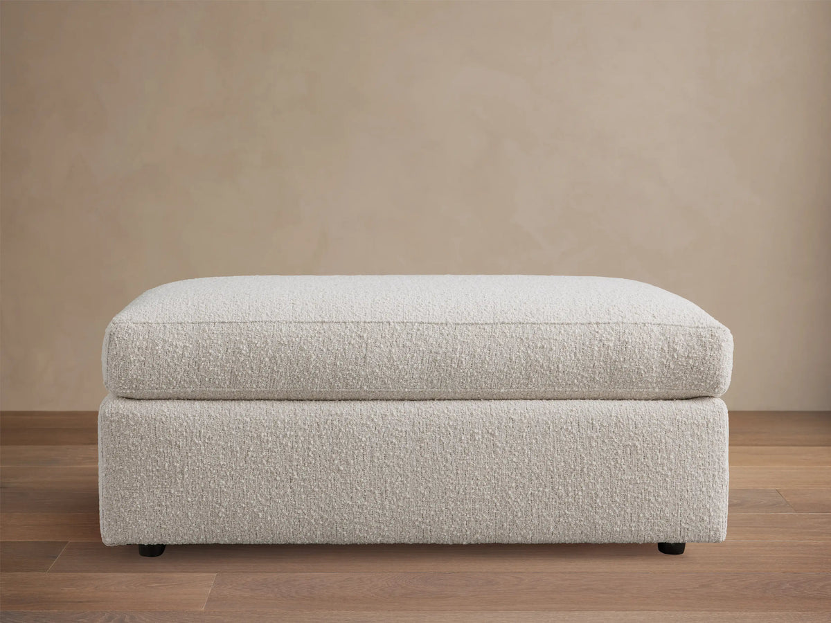 Light cream textured Vernon Ottoman, rectangular, with a soft top cushion, centered on a wooden floor.