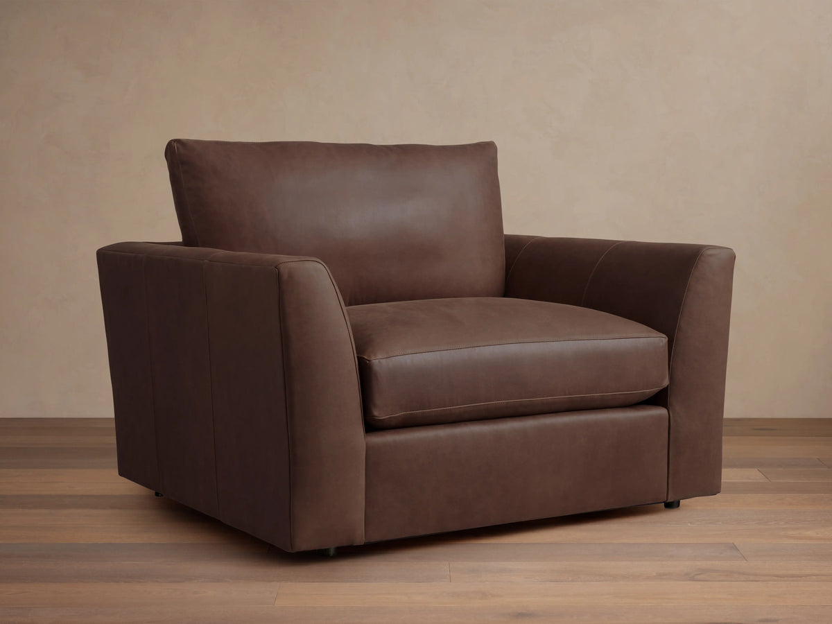 Vernon Leather Chair in dark brown leather, featuring a boxy silhouette with plush cushions and straight armrests, set against a wood floor.