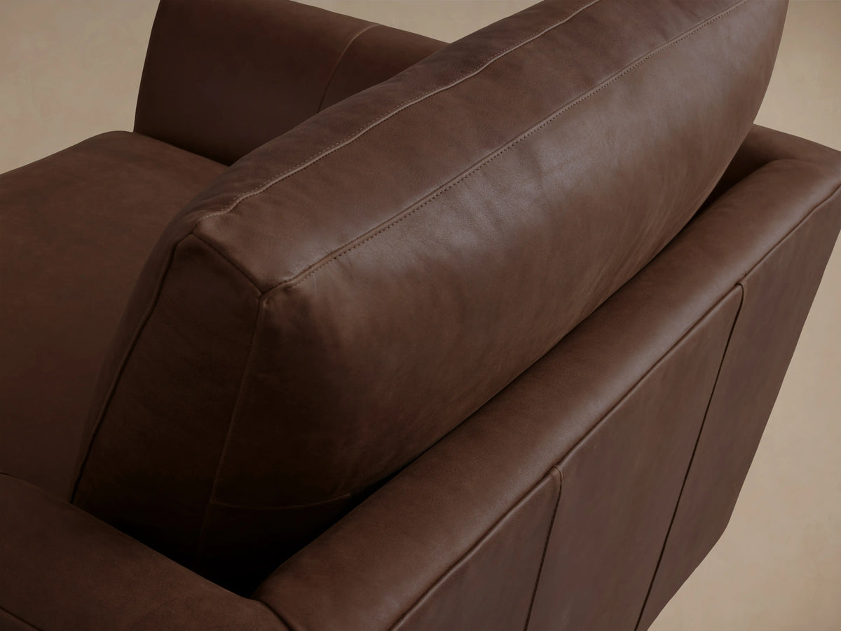 Vernon Leather Chair in rich brown leather with wide, cushioned seat and backrest. Features sleek, rounded arms and sits on a wooden floor.
