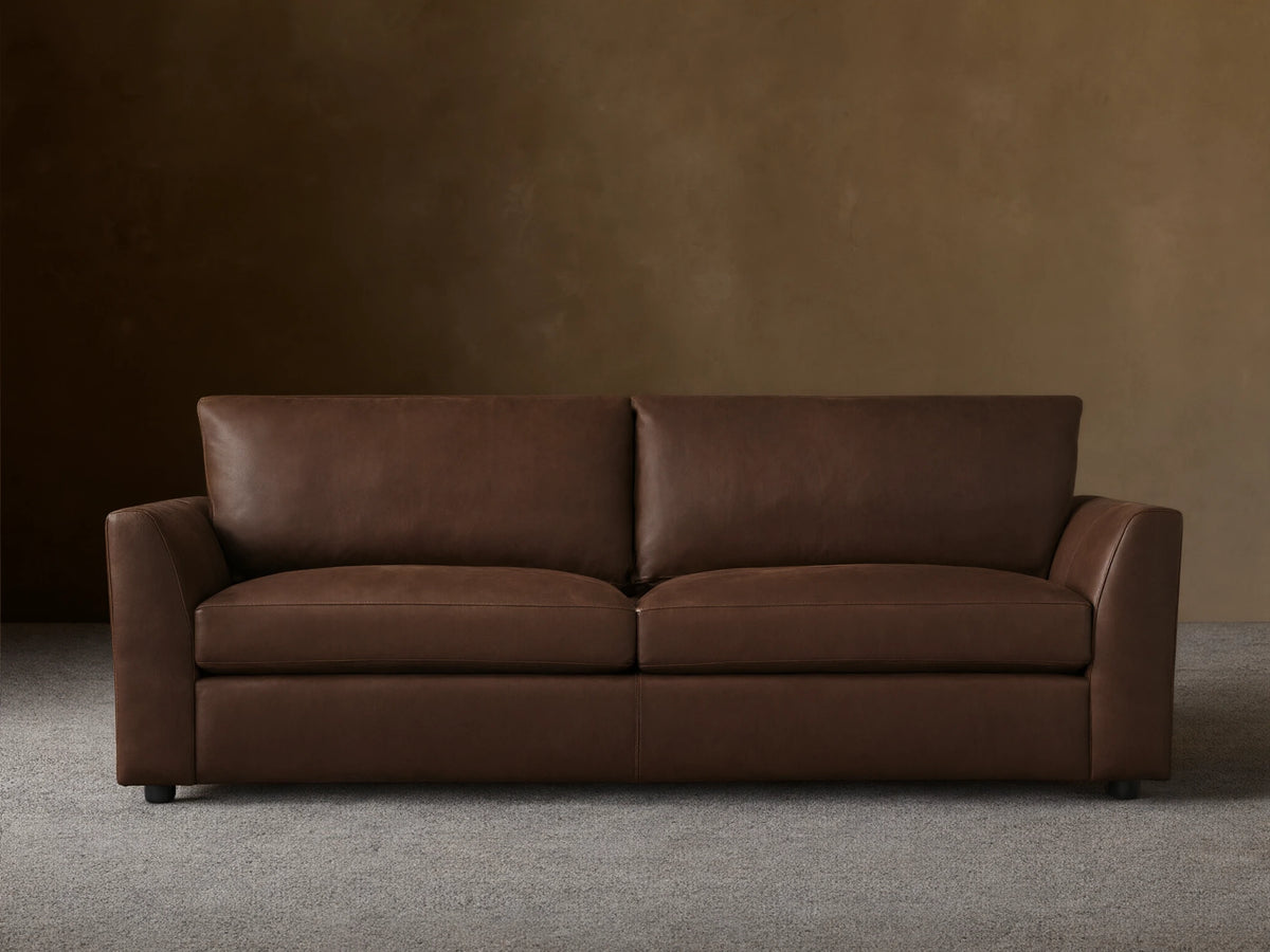 Vernon Leather Sofa featuring a rich brown finish, plush seat cushions, wide armrests, and clean lines, set against a neutral wall.