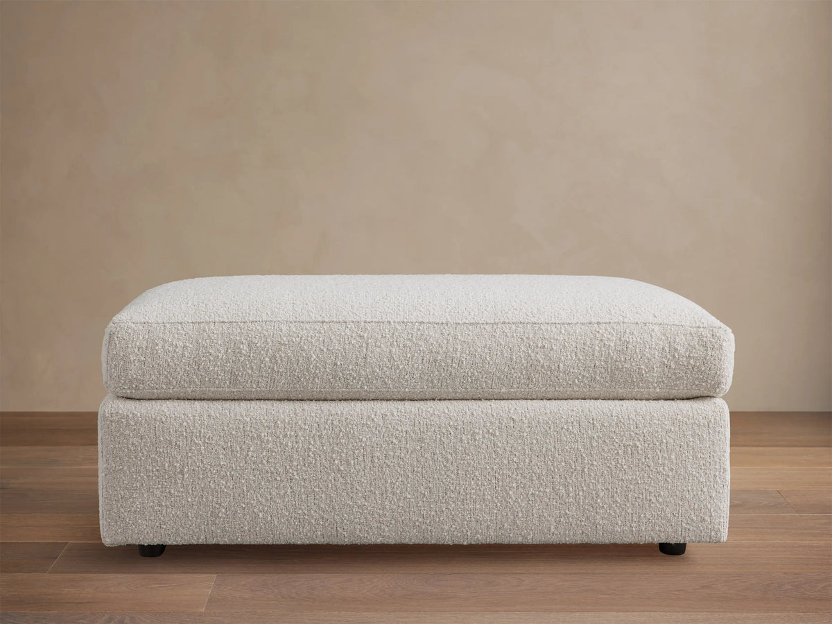 Off-white textured Vernon Square Ottoman, two-part design with plush cushion & sturdy base, on wood floor.