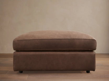 Vernon Leather Ottoman in rich brown leather, rectangular shape, flat top cushion, and low legs, set on wooden floor.