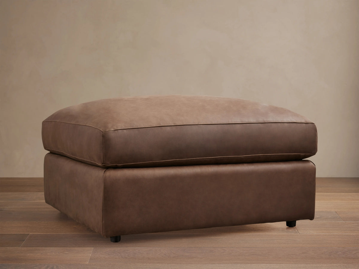 Vernon Leather Ottoman in rich brown leather on a hardwood floor, featuring a plush cushioned top and sturdy base.
