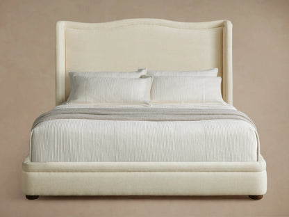 Winslet Bed with a high, curved wingback headboard and upholstered frame in light cream, white bedding, and a grey throw.