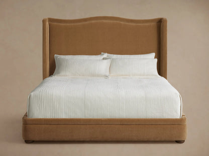 Full view of the brown upholstered Winslet Bed with a tall headboard, white bedding, and a brown throw in a sunny bedroom.