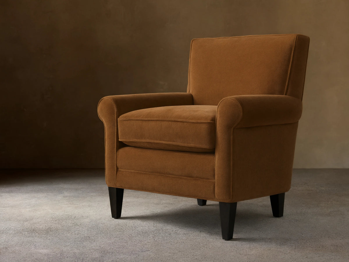 Rudy Rolled Arm Chair in cognac velvet with rolled arms, plush seat, and tapered dark legs, set against an elegant backdrop.