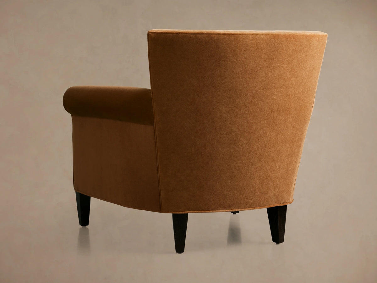 Rudy Rolled Arm Chair in cognac velvet with rolled arms, plush seat, and tapered dark legs, set against an elegant backdrop.
