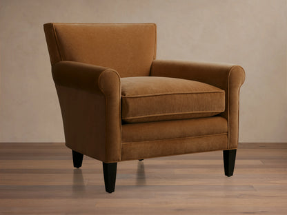 Rudy Rolled Arm Chair in cognac velvet with cushioned seat and backrest, rolled arms, and tapered legs on a wooden floor.