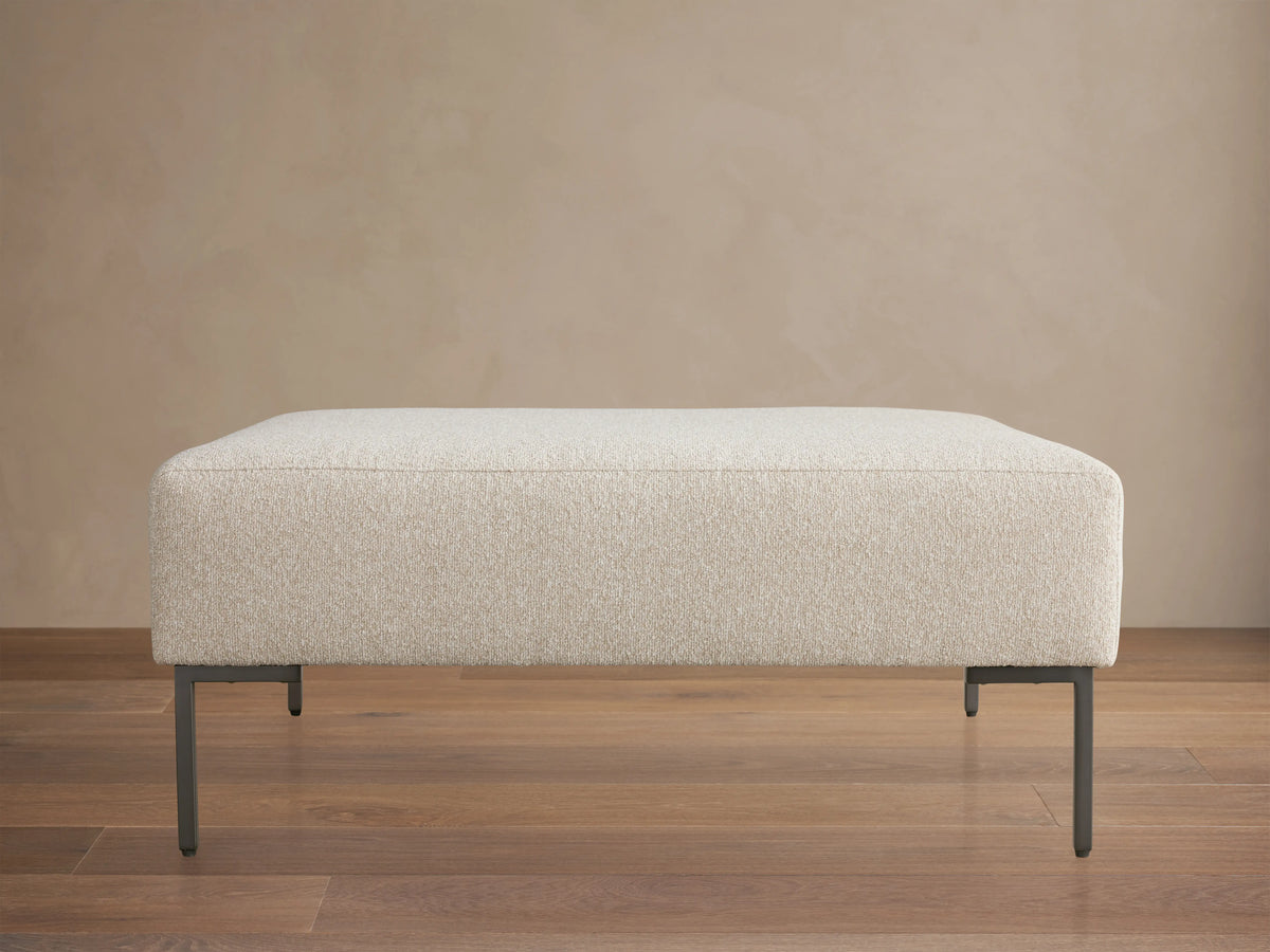 The Tilton Ottoman features a beige upholstered top with clean lines, supported by sleek black metal legs, set on a wooden floor.