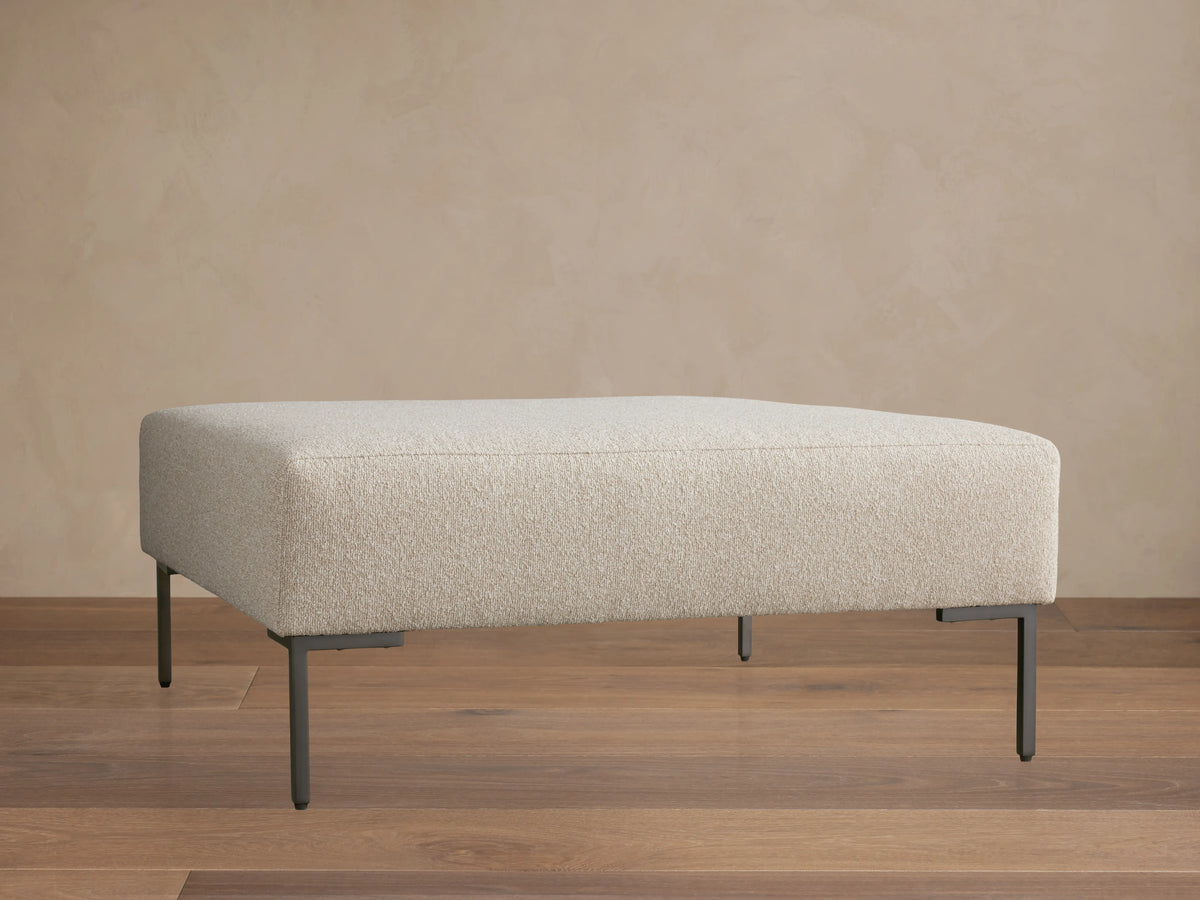 The Tilton Ottoman features a beige upholstered top with clean lines, supported by sleek black metal legs, set on a wooden floor.