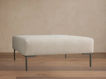 The Tilton Ottoman features a beige upholstered top with clean lines, supported by sleek black metal legs, set on a wooden floor.