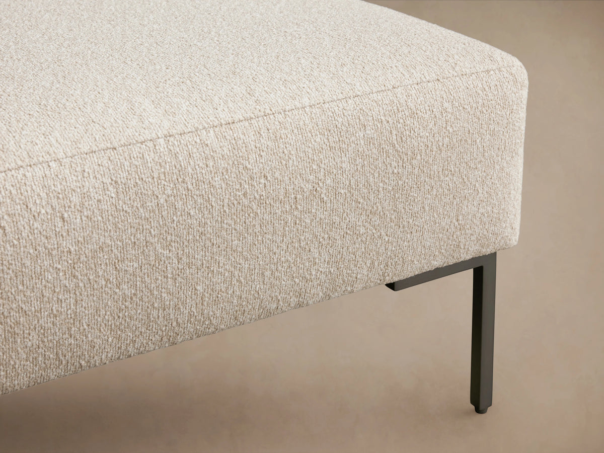 Tilton Ottoman in beige fabric with a rectangular form, set on slim, dark metal legs. Positioned on wooden floor against a beige background.