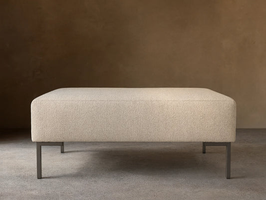 The Tilton Ottoman features a beige upholstered top with clean lines, supported by sleek black metal legs, set on a wooden floor.