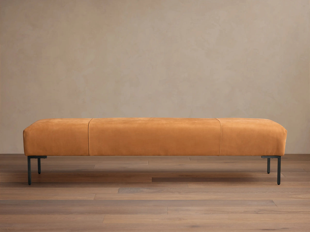 Tilton Leather Bench, long caramel leather bench with rounded ends, two seams, and black metal legs on a wood floor.