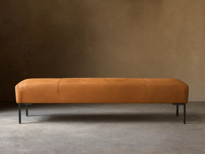 Tilton Leather Bench, long caramel leather bench with rounded ends, two seams, and black metal legs on a wood floor.