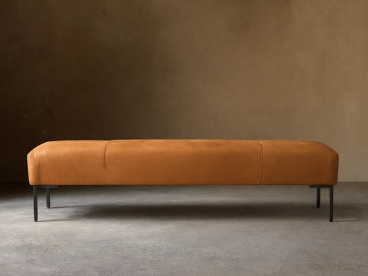 Tilton Leather Bench, long caramel leather bench with rounded ends, two seams, and black metal legs on a wood floor.