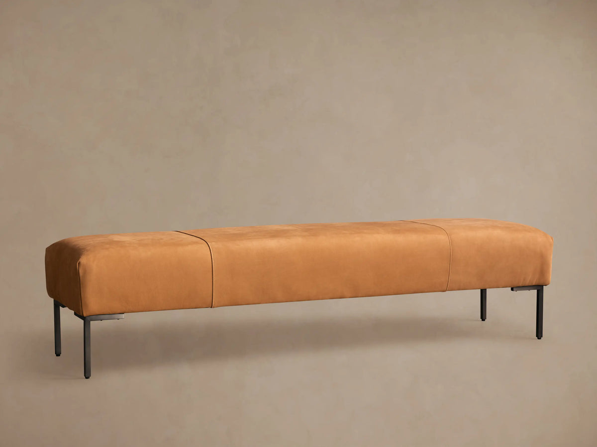 Tilton Leather Bench, long caramel leather bench with rounded ends, two seams, and black metal legs on a wood floor.
