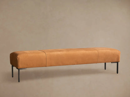 Tilton Leather Bench, long caramel leather bench with rounded ends, two seams, and black metal legs on a wood floor.