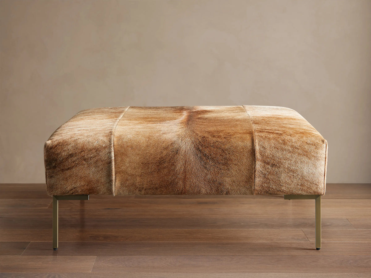 Tilton Hair on Hide Ottoman, rectangular, upholstered in blonde brindle hair-on-hide with gold metal legs, on wood floor.