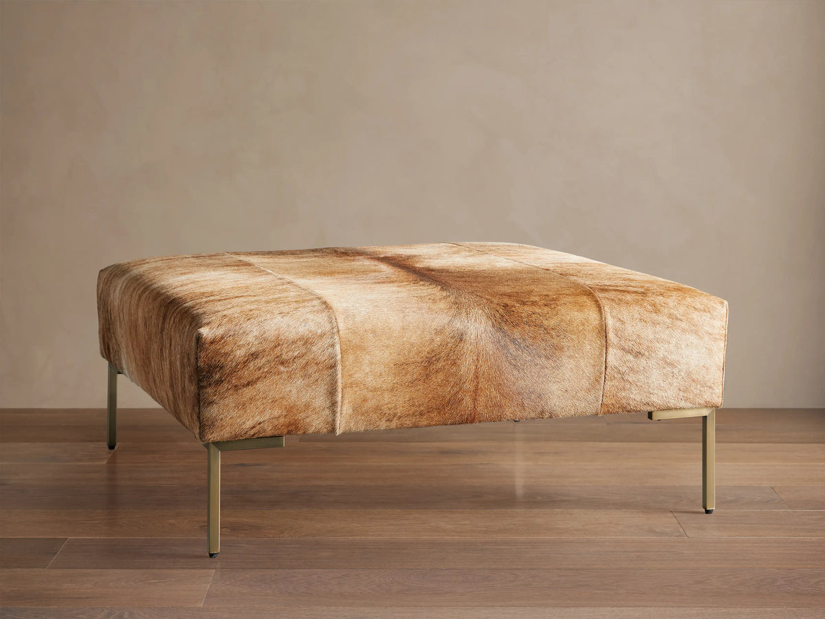 Tilton Hair on Hide Ottoman, rectangular, upholstered in blonde brindle hair-on-hide with gold metal legs, on wood floor.